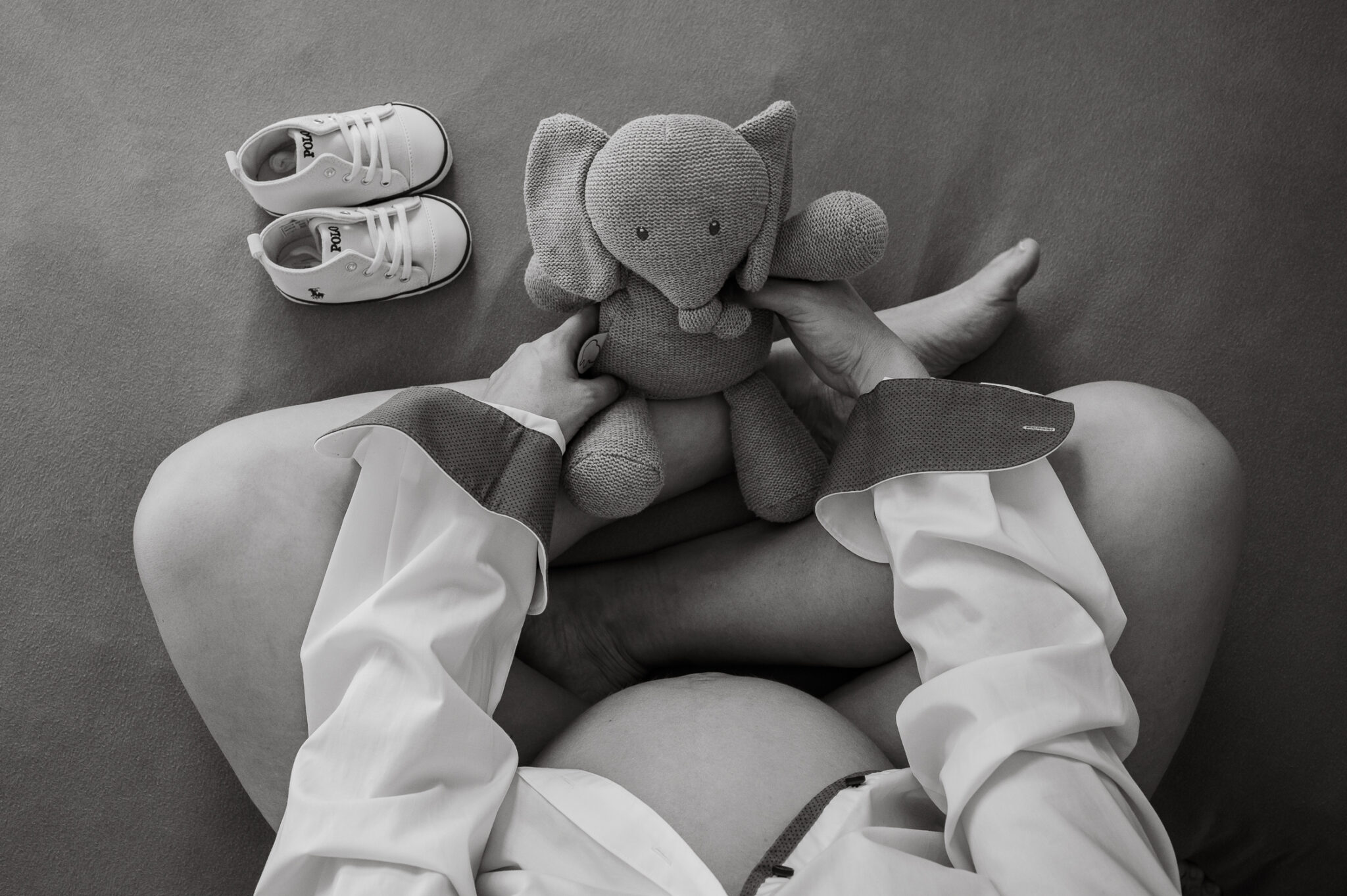 Schwangere Frau sitzt mit Kuscheltier und Babychuhen auf dem Bett