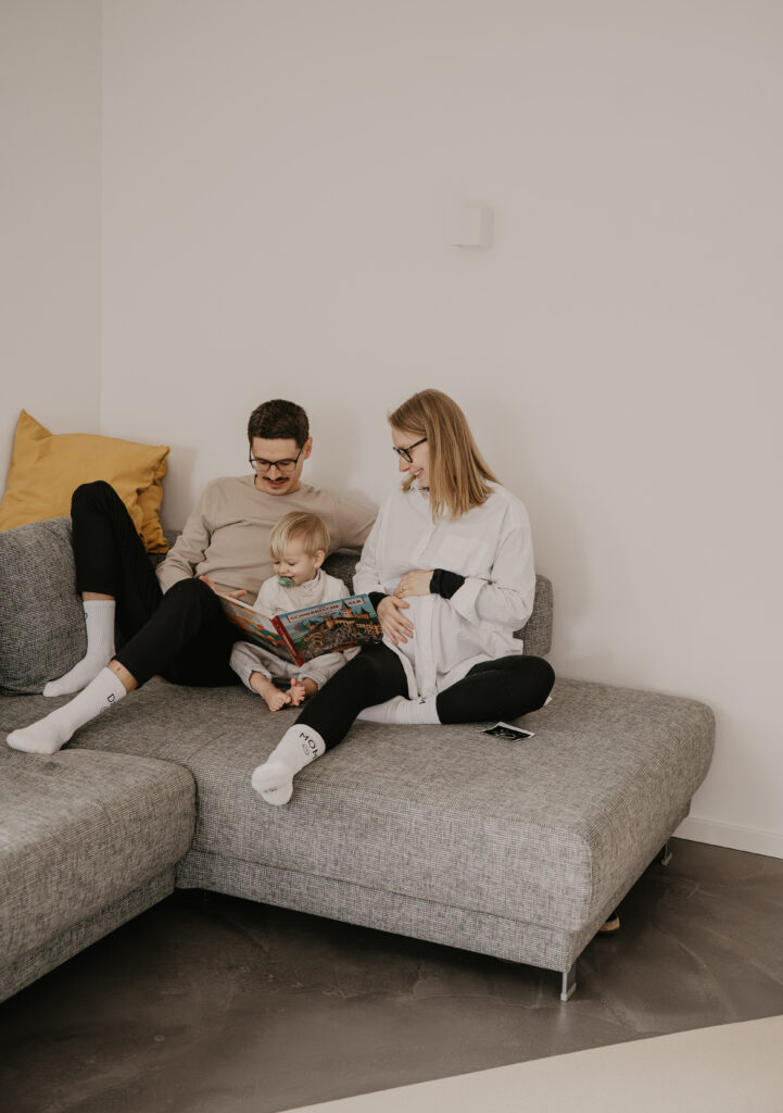Werdende Mama, Geschwisterkind und Papa sitzen auf dem Sofa und schauen sich gemeinsam ein Buch an