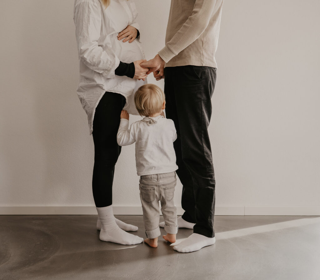 Geschwisterkind kuschelt mit dem Babybauch seiner schwangeren Mama. Frau und Mann halten Händchen