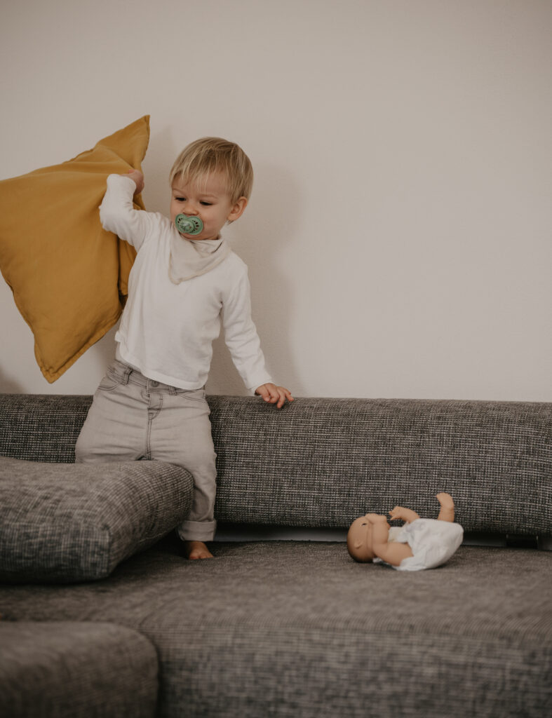 Ein kleiner Junge mit Schnuller im Mund hält ein Kissen in der Hand während er auf dem Sofa steht