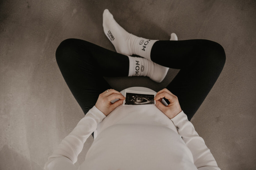 Eine schwangere Frau sitzt auf dem Boden und hält ihr Ultraschallbild vor ihren Babybauch